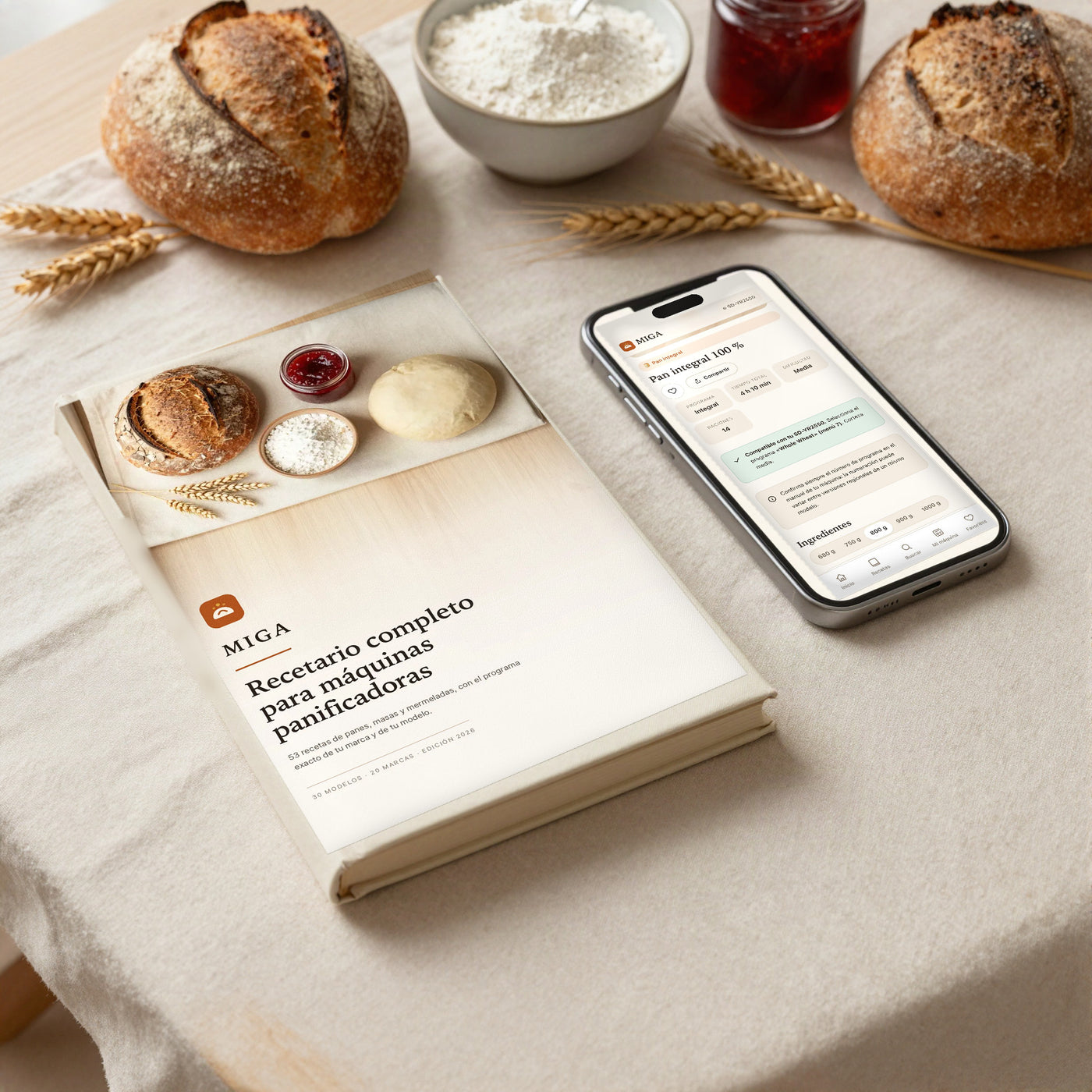 Recetario MIGA en tapa dura junto a un móvil con la mini app abierta en la receta de pan integral, sobre mantel de lino con panes, harina y mermelada.